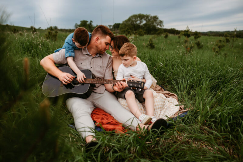Family of four sitting in a green field while the father plays the guitar