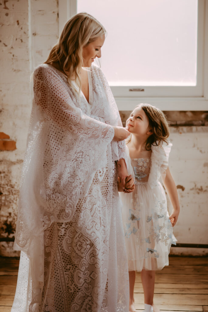 Young girl looking up at her mother while her mother holds her chin. Both are wearing long white lace dresses. 