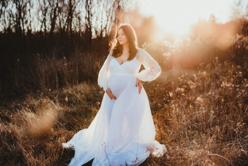 Pregnant woman standing in a field at sunset wearing a white dress
